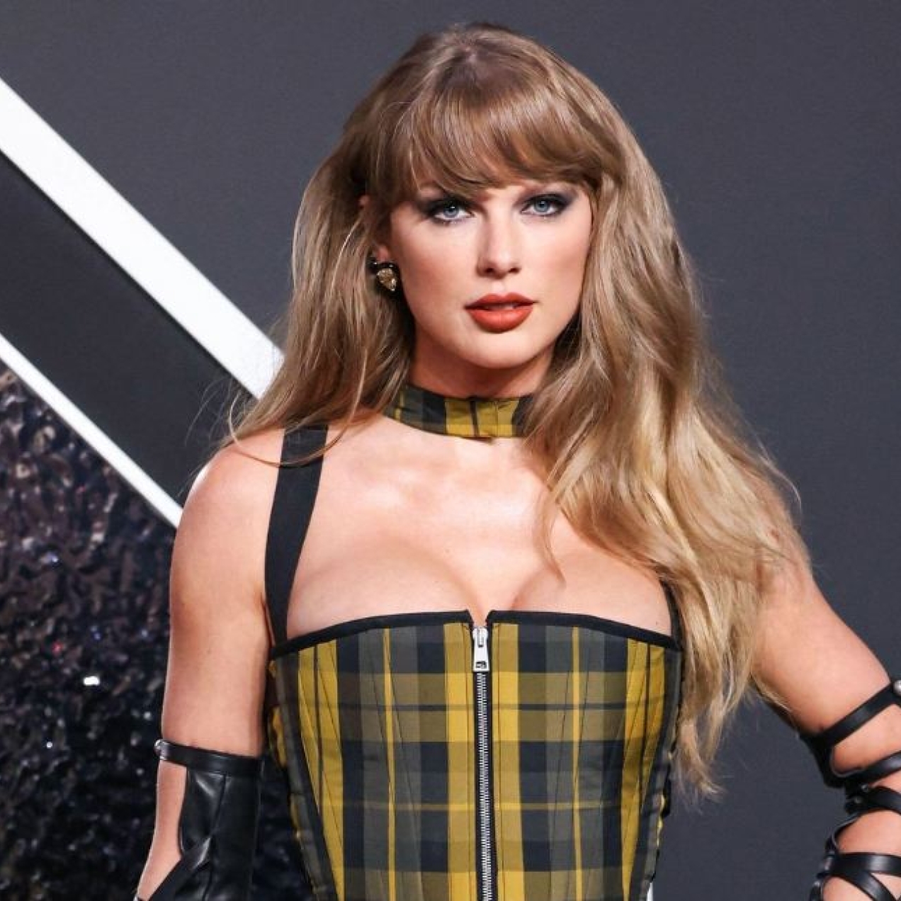 Taylor Swift poses for photographers while wearing a yellow and black check dress and black leather arm bands. The background is mostly black.